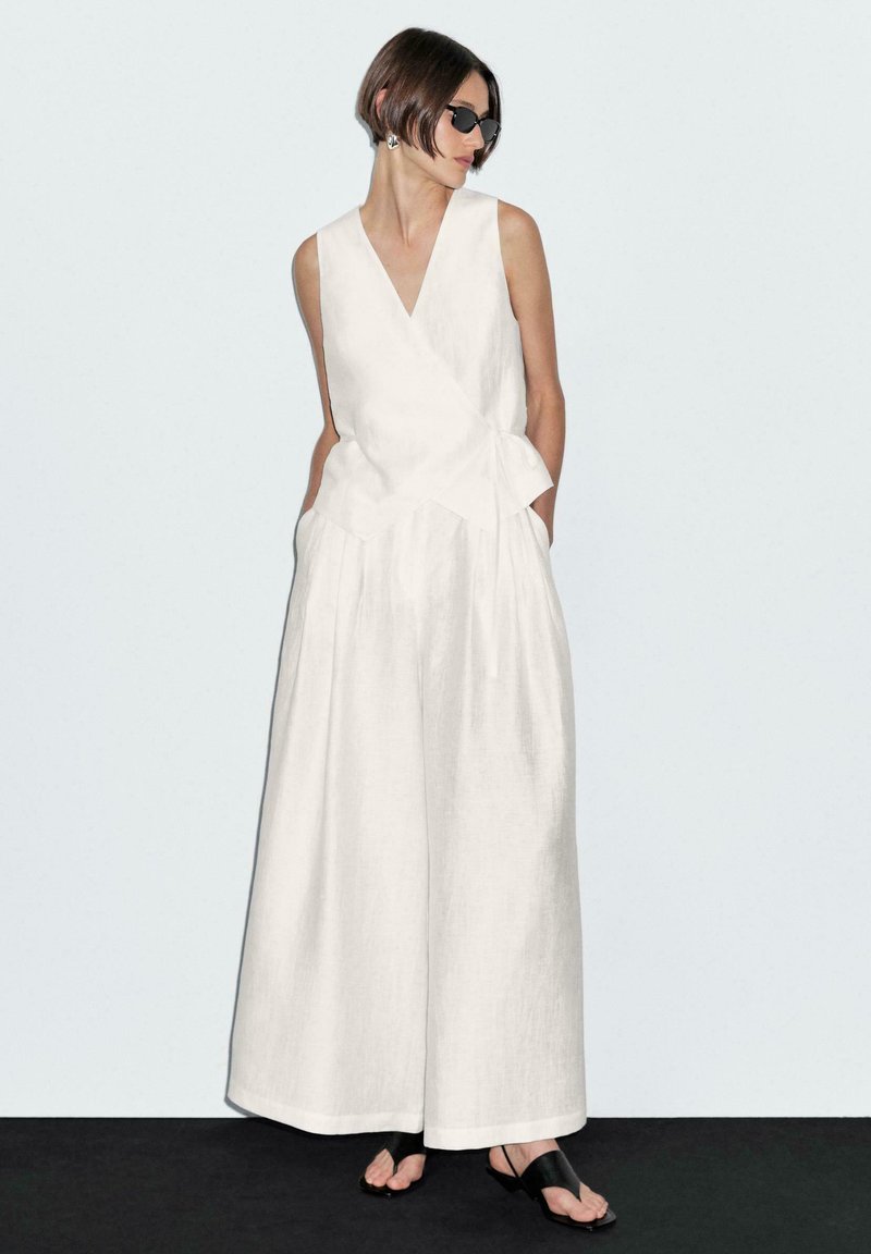 Witte linnen jumpsuit met een wikkelontwerp, brede benen en een ceintuur in de taille. Gestyled met zwarte zonnebrillen en sandalen. Soepele textuur.