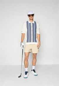 Uomo che indossa un cappello bianco, occhiali da sole, polo a righe, pantaloni beige, guanto da golf bianco e scarpe, mentre tiene un bastone da golf su uno sfondo semplice.