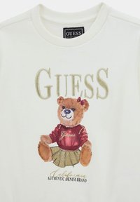 Cream sweatshirt featuring an embroidered teddy bear in a red top and glittery gold skirt, with "GUESS" text above and logo below.