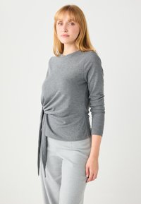 Gray long-sleeve top with a twisted front detail. Soft, lightweight fabric, crew neckline, and fitted silhouette. Paired with light gray trousers.