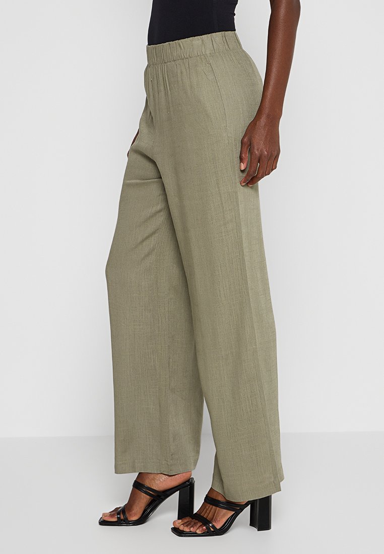 Weite, olivgrüne Hosen aus einem strukturierten Stoff. Mit einem elastischen Taillenbund ausgestattet und zu schwarzen Riemchen-Absätzen kombiniert.