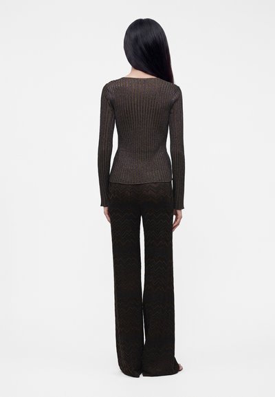 Femme aux longs cheveux noirs, vue de dos, portant un haut sombre à manches longues côtelé et un pantalon large à motifs en zigzag.