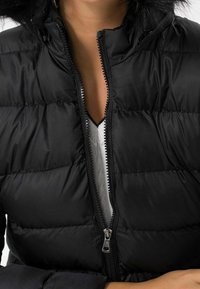 Sort pufferjakke med lynlås fortil, med hætte og horisontal quiltet mønster. Glat tekstur og detaljeret syning.
