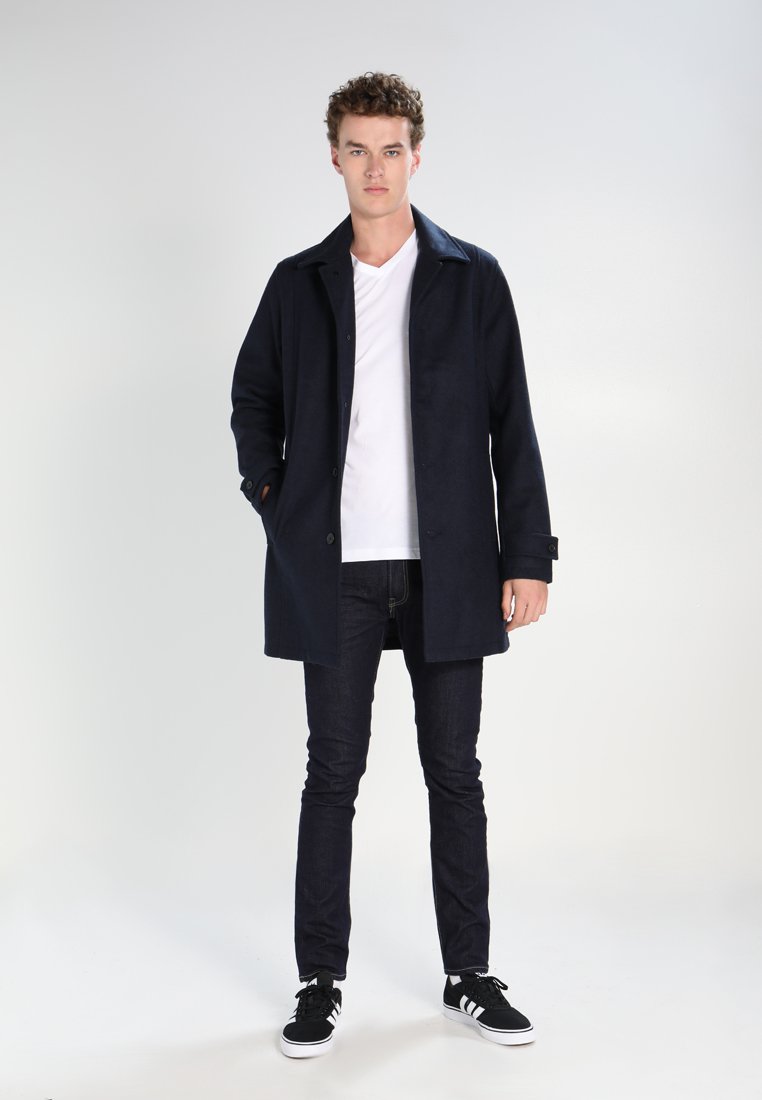 Manteau bleu marine avec un devant boutonné, un large col et des poches ; porté sur un t-shirt blanc et associé à un jean foncé et des baskets noires.