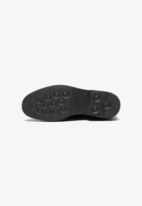 Suela de zapato de goma negra con patrones de agarre circulares y texturizados, forma ligeramente curva con un borde suave y nombre de marca en relieve.