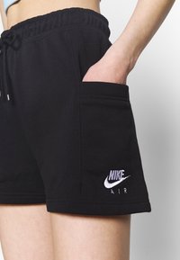 Hand i fickan på svarta Nike Air-shorts med dragsko i midjan, bärs på nakna ben mot en enfärgad bakgrund.