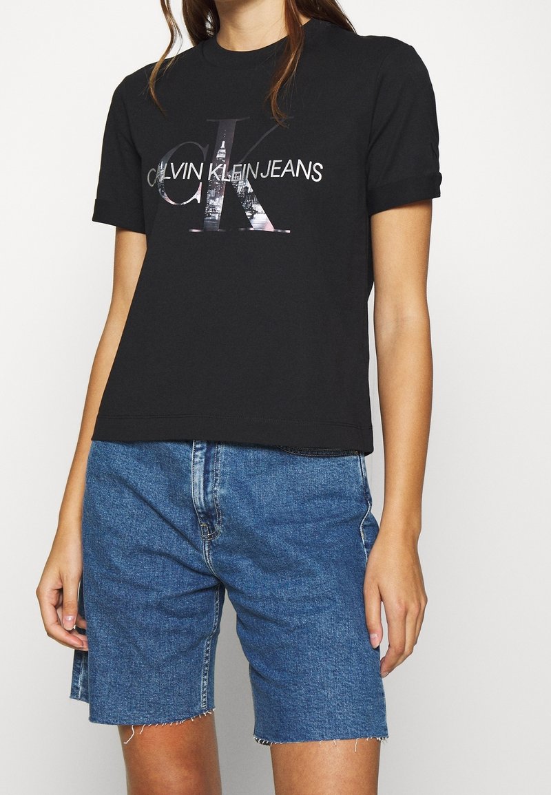 Black cotton t-shirt featuring a large "CK" graphic with the text "CALVIN KLEIN JEANS" beneath, paired with blue denim shorts with frayed edges.