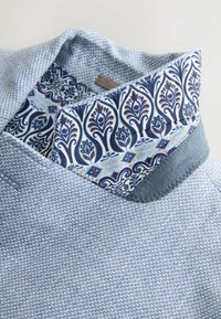 Lichtblauwe geweven jaskraag omgeslagen, met zichtbare binnenvoering met een blauw-wit bloemdecoratief patroon.