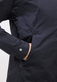 Κοντινό πλάνο μαύρου μπουφάν που δείχνει ένα χέρι να έχει εισαχθεί σε πλαϊνή τσέπη, με ένα μικρό λευκό λογότυπο στην μανσέτα του μανικιού.