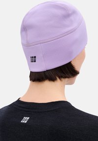 Lila Beanie mit strukturierter Oberfläche, mit einem kleinen schwarzen Logo auf der Rückseite. Die Person trägt ein schwarzes Shirt mit einem ähnlichen Logo.