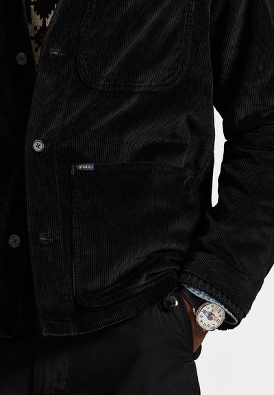 Black corduroy jacket features a front pocket, button closure, and a Polo tag. A silver watch with a colorful face peeks from beneath.