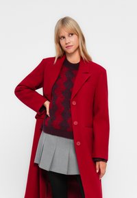 Red wool blend coat with a relaxed fit, single-breasted design, and large buttons. Worn over a patterned sweater and grey pleated skirt.