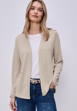 Donna bionda che indossa un cardigan beige aperto sul davanti, una maglietta bianca, jeans blu e una cintura con stampa leopardata, posando con una mano in tasca.
