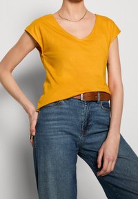 Mosterdgele katoenen t-shirt met een v-hals en korte mouwen, gecombineerd met hooggetailleerde blauwe spijkerbroek en een bruine leren riem.