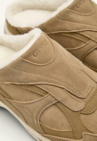 Beige színű, puhatalpú, nyúlszőr béléssel ellátott mokaszin cipők. Réteges kialakítás jellemzi, varrott díszítésekkel és texturált felülettel a jobb tapadás érdekében.