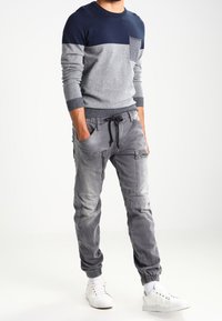 Maglione a blocchi di colore grigio e blu navy con scollo tondo e taschino sul petto, abbinato a pantaloni cargo grigi con tasche e polsini elasticizzati. Sneakers bianche.