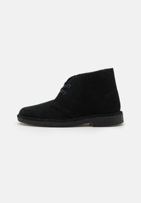 Schwarze Wildleder-Stiefelette mit runder Zehenpartie, zwei Schnürlöchern an der Seite und einer strukturierten Gummisohle. Minimale Nähte.