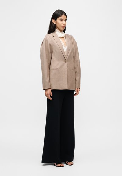 Blazer beige oversize avec épaules structurées, fermeture à un bouton, deux poches avant ; associé à un pantalon large noir et des sandales.