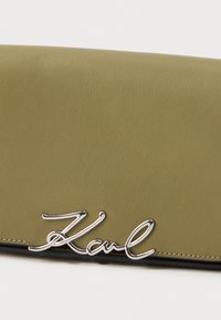 Olivadgrön läderplånbok med silverfärgad metalldetalj med "Karl" signatur på den främre nederkanten.