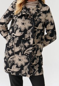 Abrigo de patrón floral en negro y beige, hecho de una tela texturizada. Presenta un cuello y bolsillos laterales, con un corte de silueta relajada.