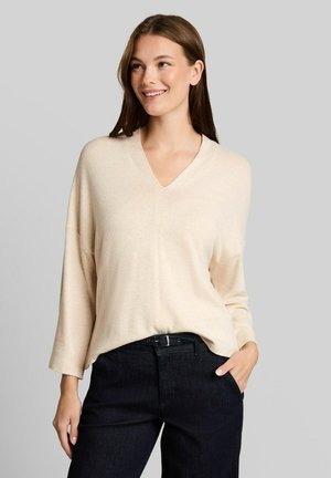 Jonge vrouw die lacht, draagt een losse beige V-hals trui en zwarte broek met haar linkerhand in de zak, tegen een effen grijze achtergrond.