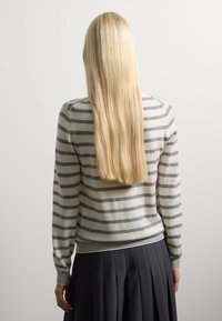 Personne avec de longs cheveux blonds raides portant un pull rayé gris et blanc et une jupe plissée noire, se tenant contre un mur uni.