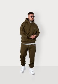 Olive green hoodie and sweatpants set, made of soft fabric. Features a front pocket, drawstring waist, and white sneakers. Minimalistic logo detail.