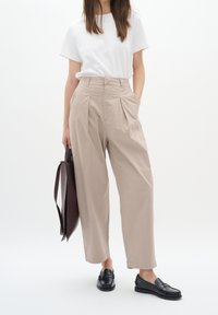 Beige plisé kalhoty s vysokým pasem, v kombinaci s bílým tričkem a černými polobotkami, drží tmavě hnědou tašku. Hladká textilie.
