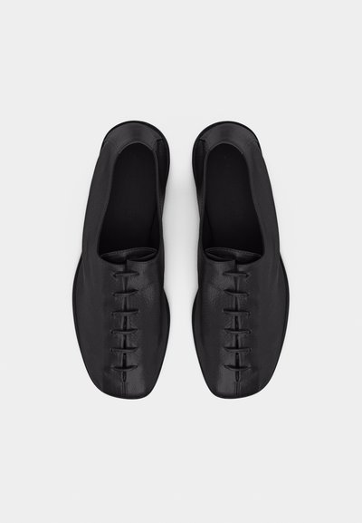Ζευγάρι μαύρα δερμάτινα μινιμαλιστικά παπούτσια με μπροστινό ελαστικό κορδόνι, στρογγυλεμένες μύτες και χαμηλές τακούνια, φωτογραφημένα από πάνω σε λευκό φόντο.