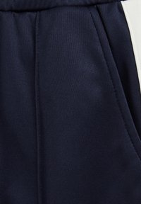 Tuvumā redzams tumši zils audums ar sānu kabatu un baltu vertikālu strīpu, uzrādot tekstūru un šuvju detaļas.