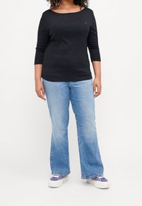 Musta pitkähihainen paita, jossa on veneenmuotoinen kaula-aukko, yhdistettynä vaaleansinisiin leveälahkeisiin farkkuihin. Jalkineissa on violetteja yksityiskohtia ja valkoinen pohja.