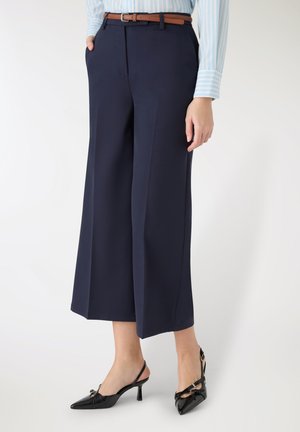 Granatowe spodnie o szerokich nogawkach z gładką fakturą i przednimi kieszeniami, zestawione z czarnymi sandałkami na obcasie z paseczkiem z tyłu, ozdobionymi kokardą. Dołączony brązowy pasek.