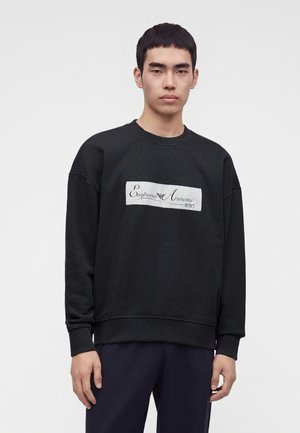 Jeune homme portant un sweat-shirt noir avec un écusson logo blanc Emporio Armani sur la poitrine, debout devant un fond clair uni.