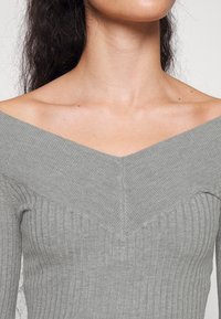 Femme aux longs cheveux foncés portant un pull gris côtelé à épaules dénudées, laissant apparaître le cou et le haut de la poitrine.