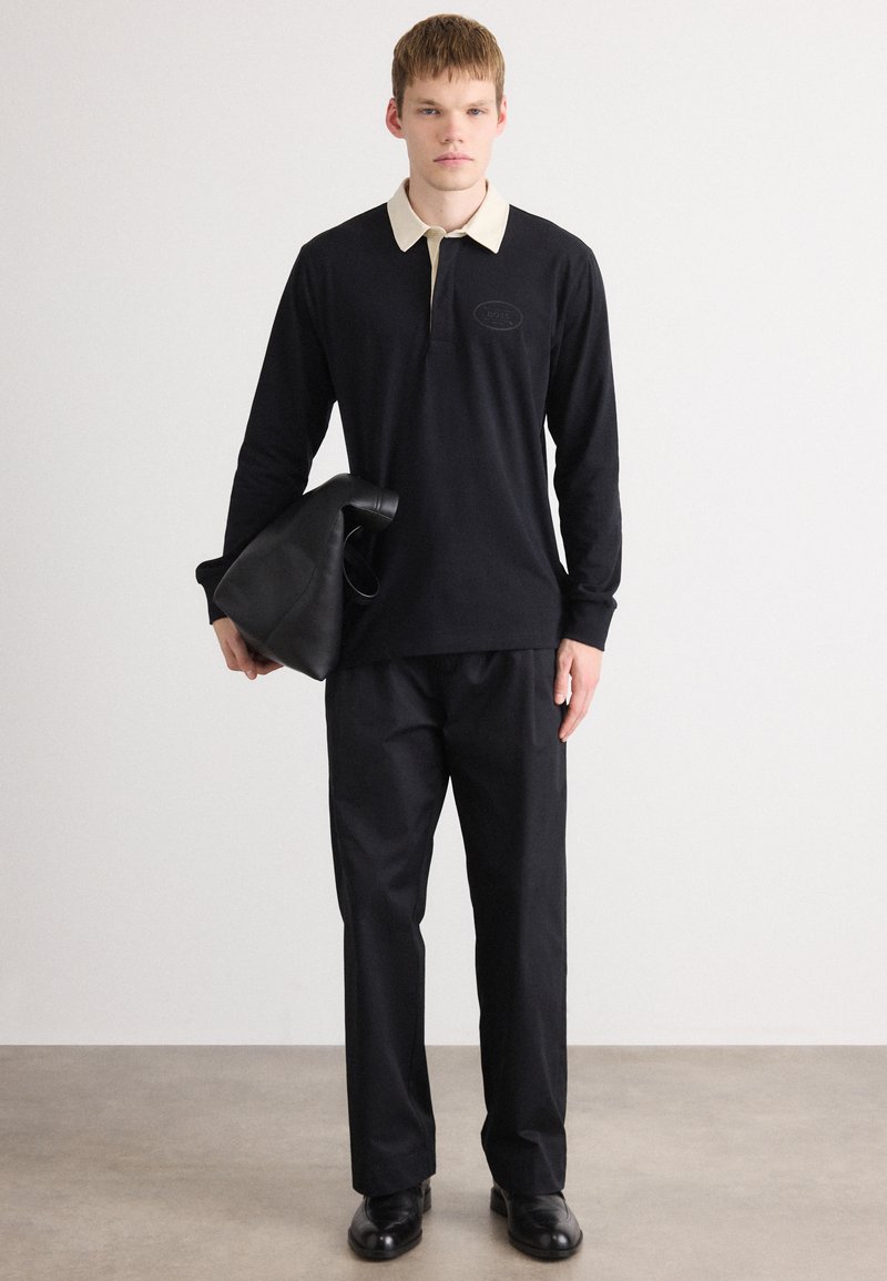 Jonge man in een zwart poloshirt met lange mouwen en een beige kraag, zwarte broek, zwarte schoenen, met een opgevouwen zwarte leren tas in zijn rechterhand.