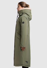 Lang olivengrøn parka med hætte foret med pels, kviltede detaljer, sidelommer og et trekantet logo-patch på kanten.