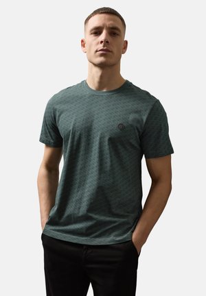 Mann mit kurzem Haar, der ein dunkelgrünes T-Shirt mit Muster und schwarze Hose trägt, steht mit den Händen in den Taschen vor einem einfarbigen Hintergrund.