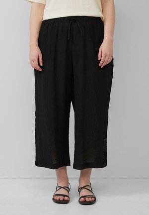 Pantalons larges noirs en tissu texturé avec une taille élastique et un cordon de serrage. Associés à des sandales noires à brides.