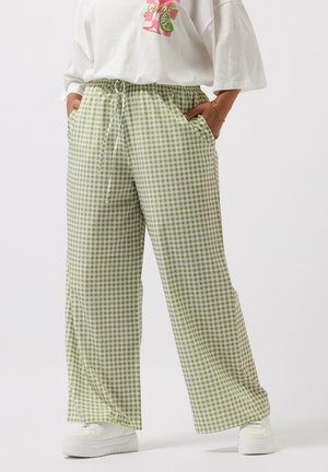 Person trägt lockere, grün-weiße Vichy-Karo-Hose mit weitem Bein und elastischem Bund sowie weißen Turnschuhen, Hände in den Taschen.