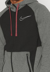Haut à capuche noir et gris avec une finition texturée. Doté de cordons rouges et de poches latérales. Logo Nike blanc sur la poitrine.