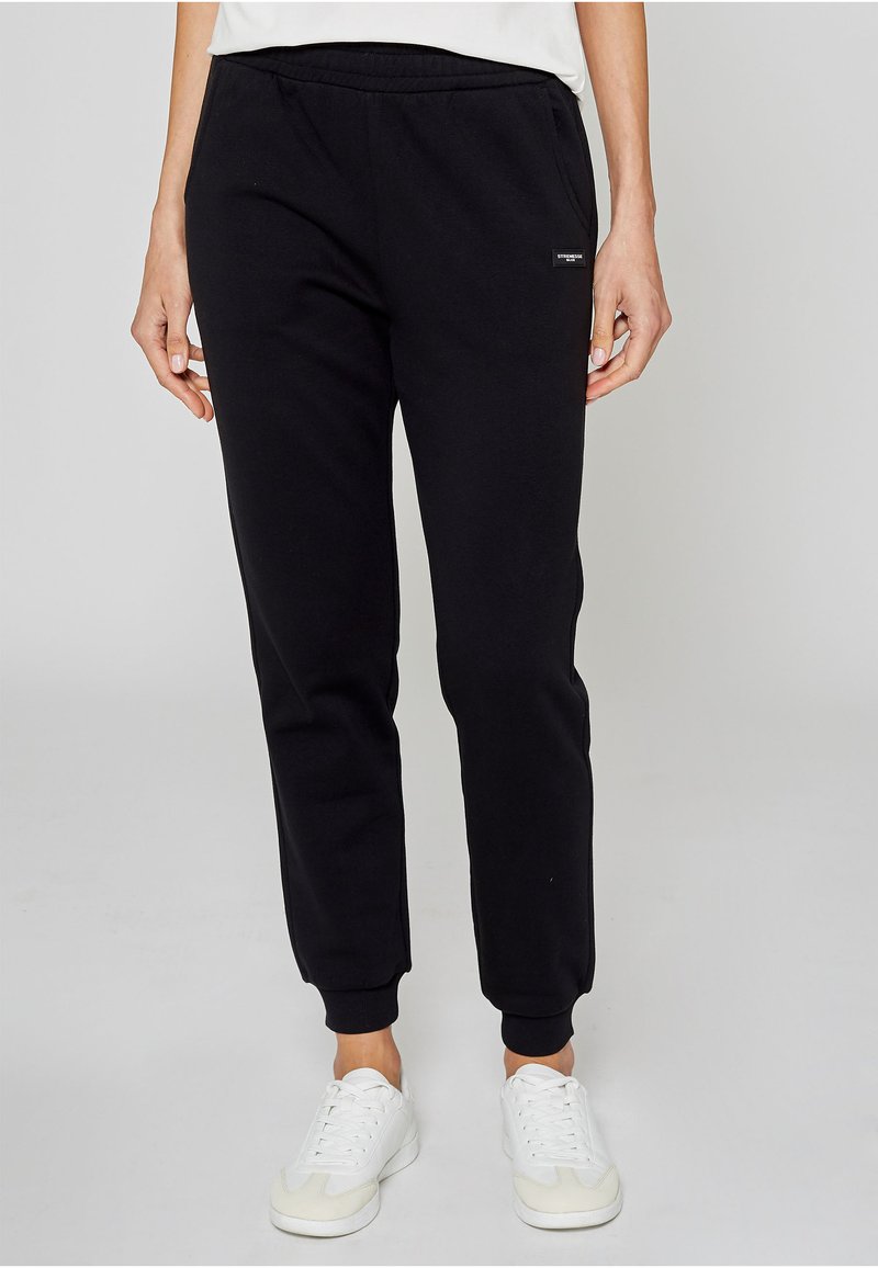 Zwarte joggingbroek van zacht materiaal, voorzien van een elastische tailleband, geribbelde boorden en een klein logoplaatje aan de zijkant.