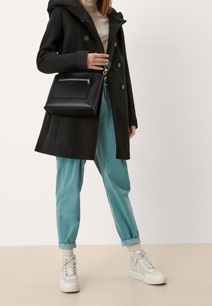 Schwarzer Wollmantel mit Kapuze, beiger Pullover und hellblaue Hose. Schwarze Ledertasche mit vorderer Reißverschlusstasche. Weiße hohe Sneakers.