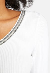 Primer plano de una persona que lleva una camiseta blanca de cuello en V con un ribete plateado y gris a lo largo del escote.