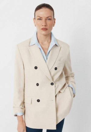 Kvinde med tilbagelagt hår iført en beige dobbeltradet blazer over en blåstribet skjorte, stående foran en ensfarvet hvid baggrund.