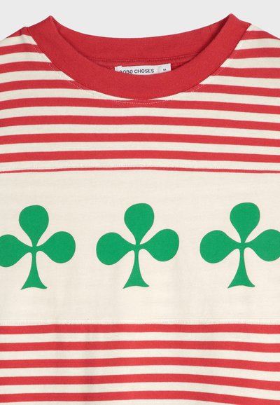 T-shirt a righe rosse e crema con tre grandi motivi di trifoglio verde centrati sul petto. Materiale in cotone morbido, design a girocollo.