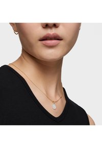 Collar de cadena de oro con un colgante ovalado texturizado. Llevado sobre un top sin mangas negro, con piel suave y labios sutiles.