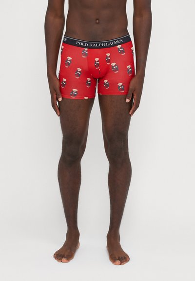 Shorts de bain rouges avec un motif de ours répété dans différentes tenues. Taille élastique avec la marque "POLO RALPH LAUREN". Texture de tissu lisse.