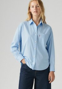 Camicia azzurra in cotone con maniche lunghe, colletto a punta e taschino sul petto con il logo del brand. Indossata con jeans scuri.