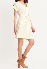 Vestido color crema con mangas cortas, escote en V, cintura ceñida con cinturón y bolsillos. Combinado con botas de tobillo marrón con volantes y tacones de bloque.