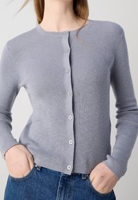 Světlounce šedý žebrovaný pletený kardigan s kulatým výstřihem, který má na přední straně šest bílých knoflíků a přiléhavé dlouhé rukávy.